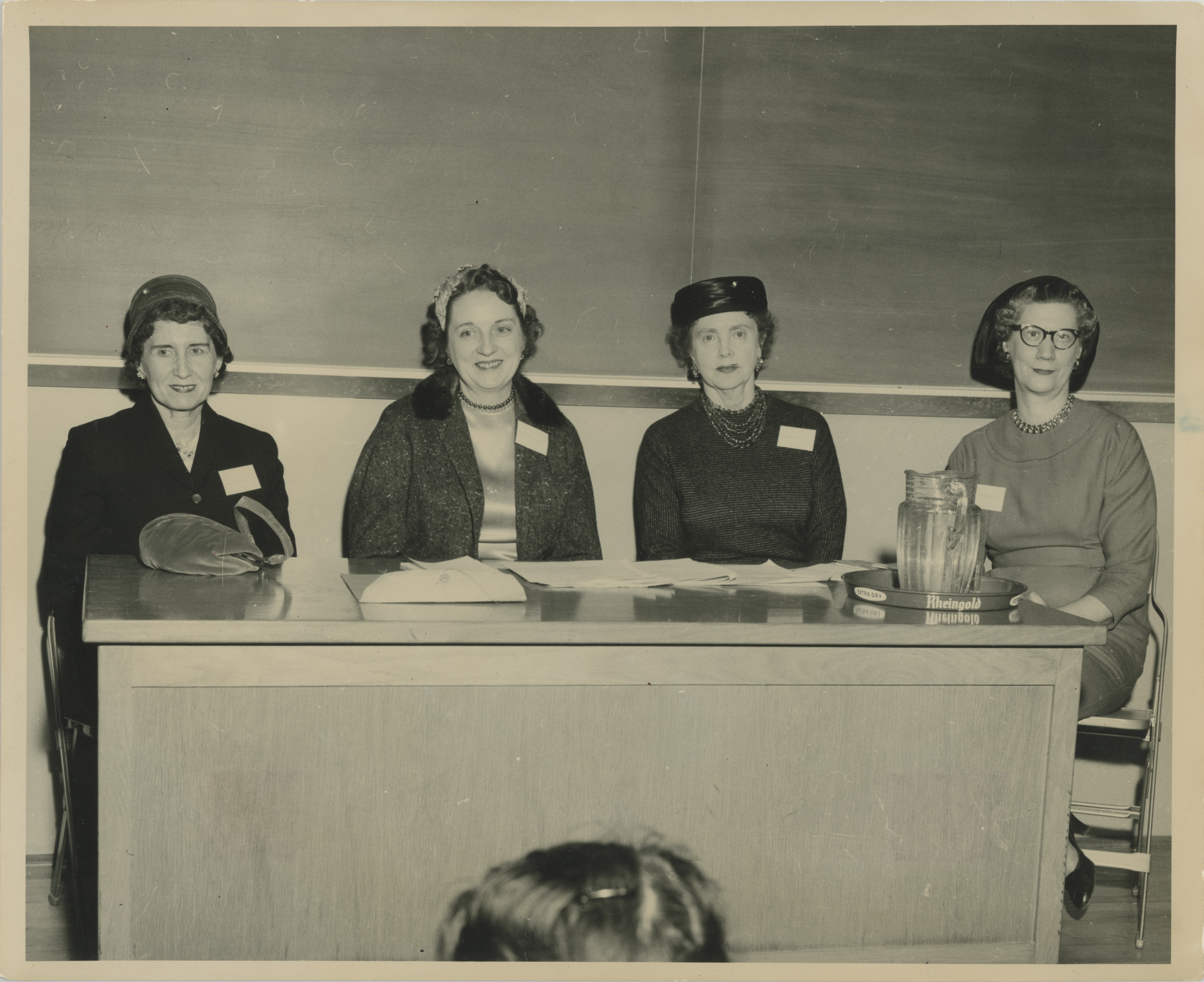 Black and white aged image of four women sitting at a desk smiling toward the camera, a chalk board in the background
