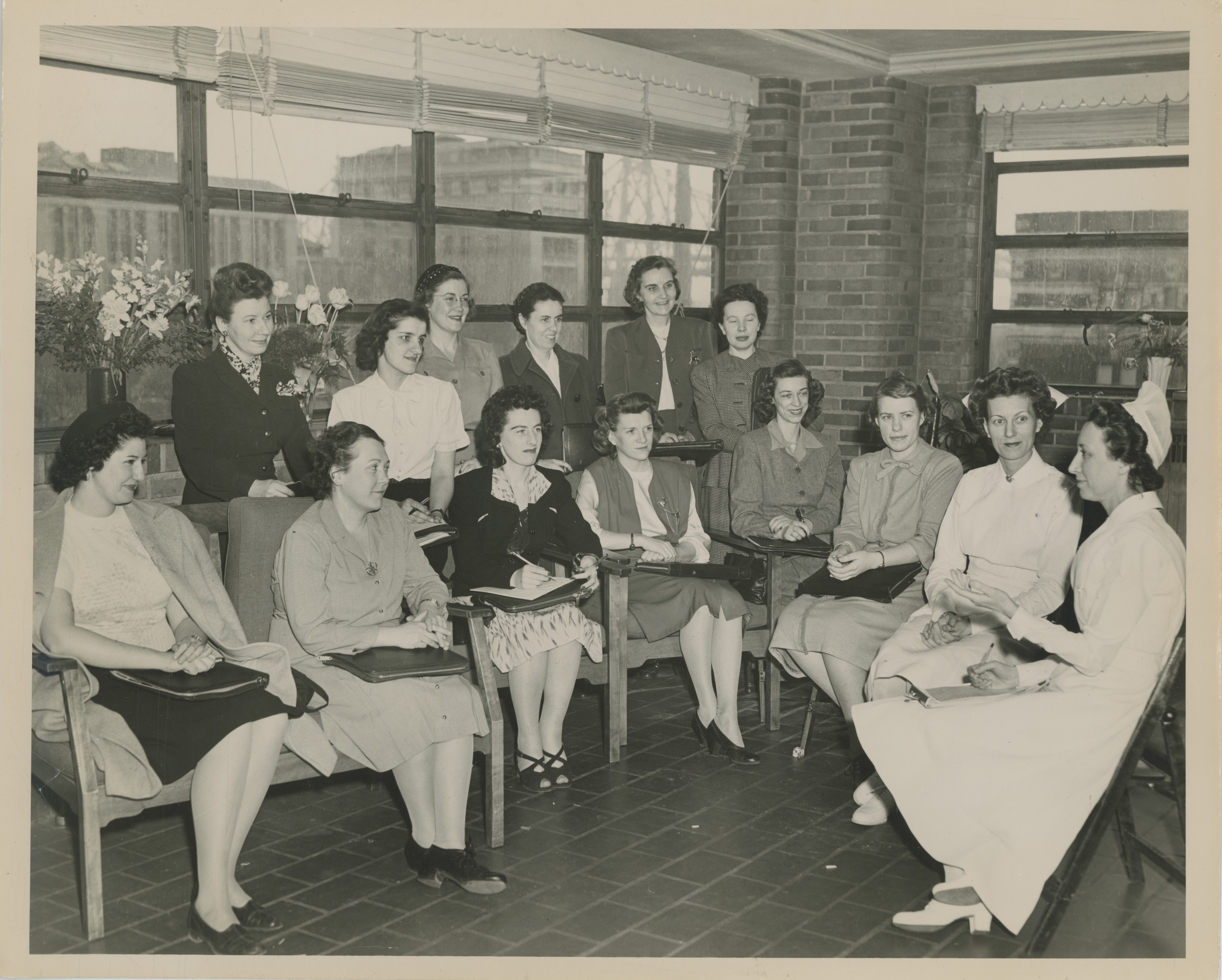 Black and white aged image of crowded room with women sitting in chairs