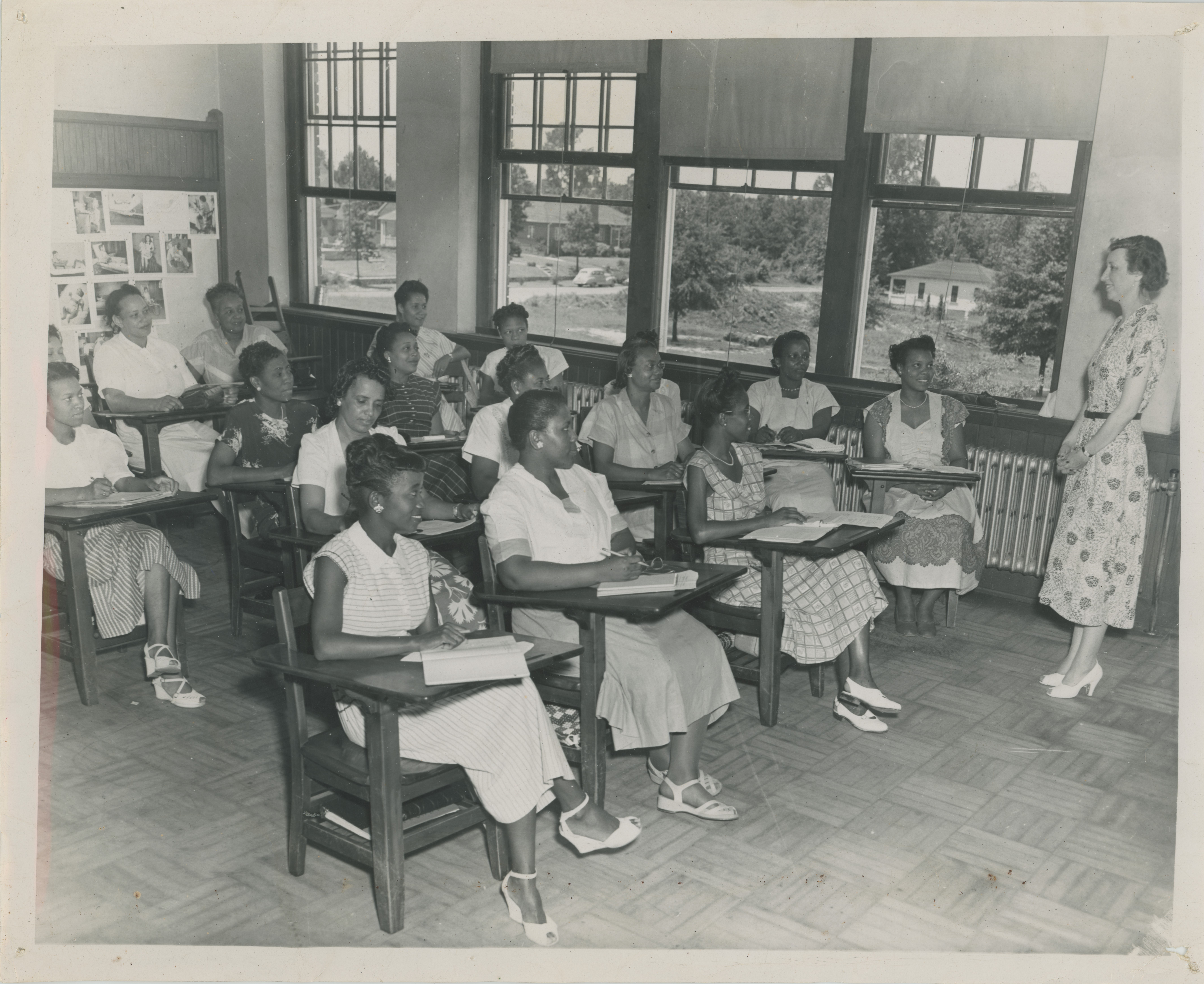 Black and white aged image of a classroom with African American female students sitting in chairs as Caucasian female, Katherine Nelson, teaches at the front of the class 